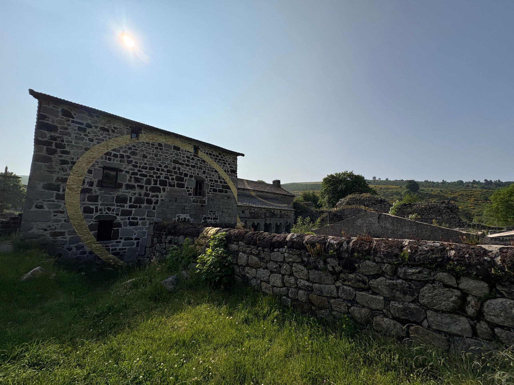 Bâtiment en pierre de l'abbaye de Mazan traversé par un grand arc de cercle doré, mur de pierres sèches au premier plan, soleil dans le ciel