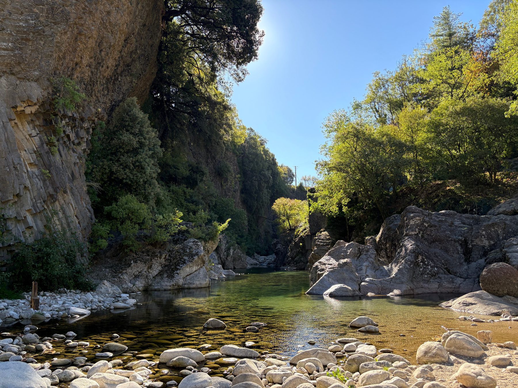 Bassin d’eau claire et verte entre rochers polis, falaise basaltique à gauche et arbres ensoleillés à droite