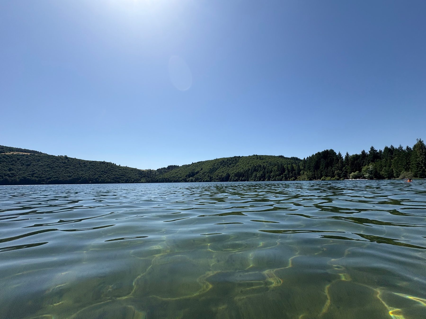 Surface du lac d’Issarlès vue au ras de l’eau, fond clair visible et rives boisées à l’horizon