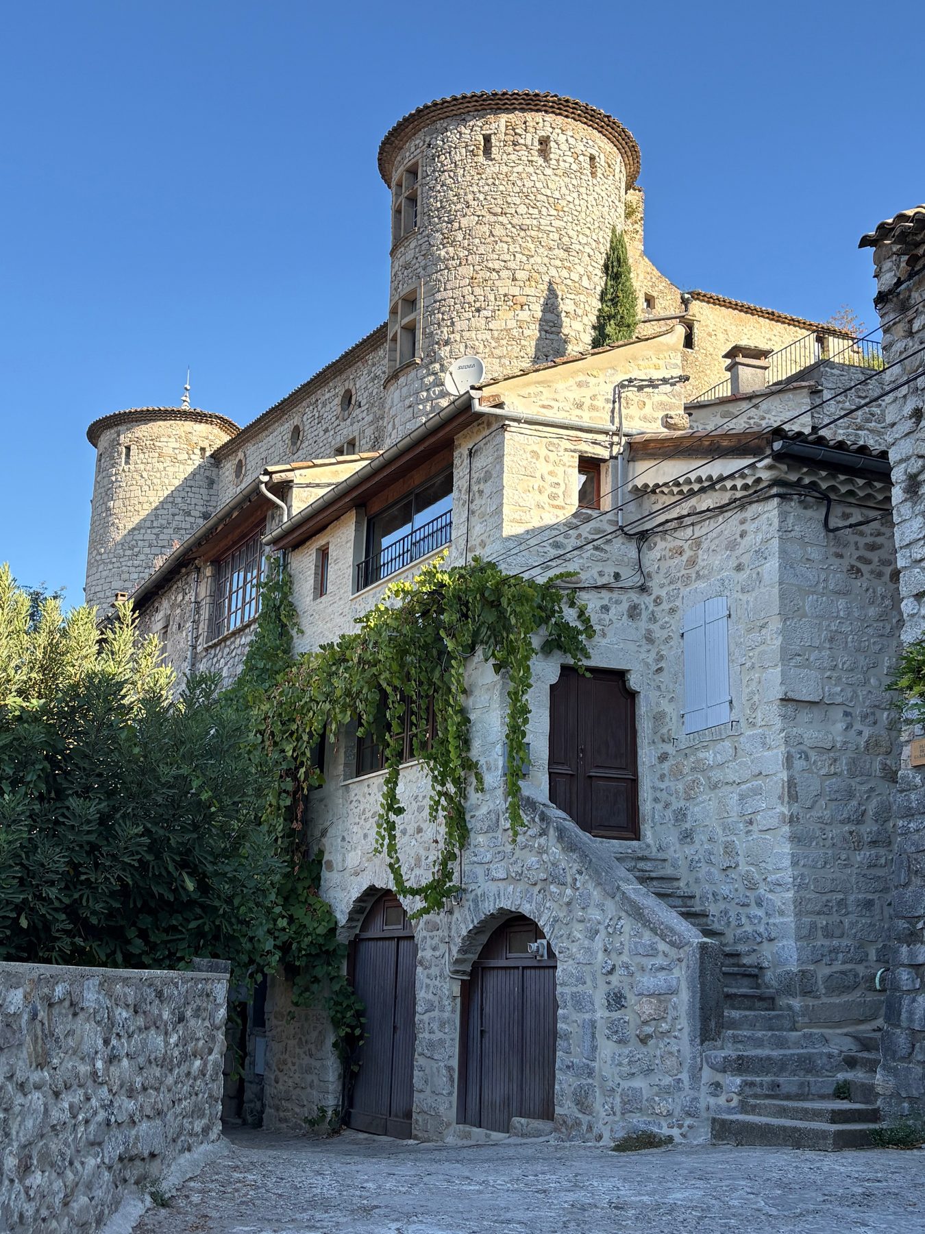 Tours de pierre du château de Vogüé dominant une maison ancienne à escalier extérieur et treille