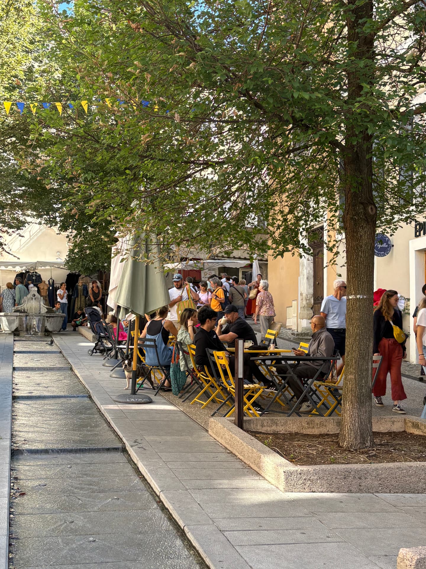 Rue ombragée avec terrasse aux chaises jaunes, fontaine et passants un jour de marché