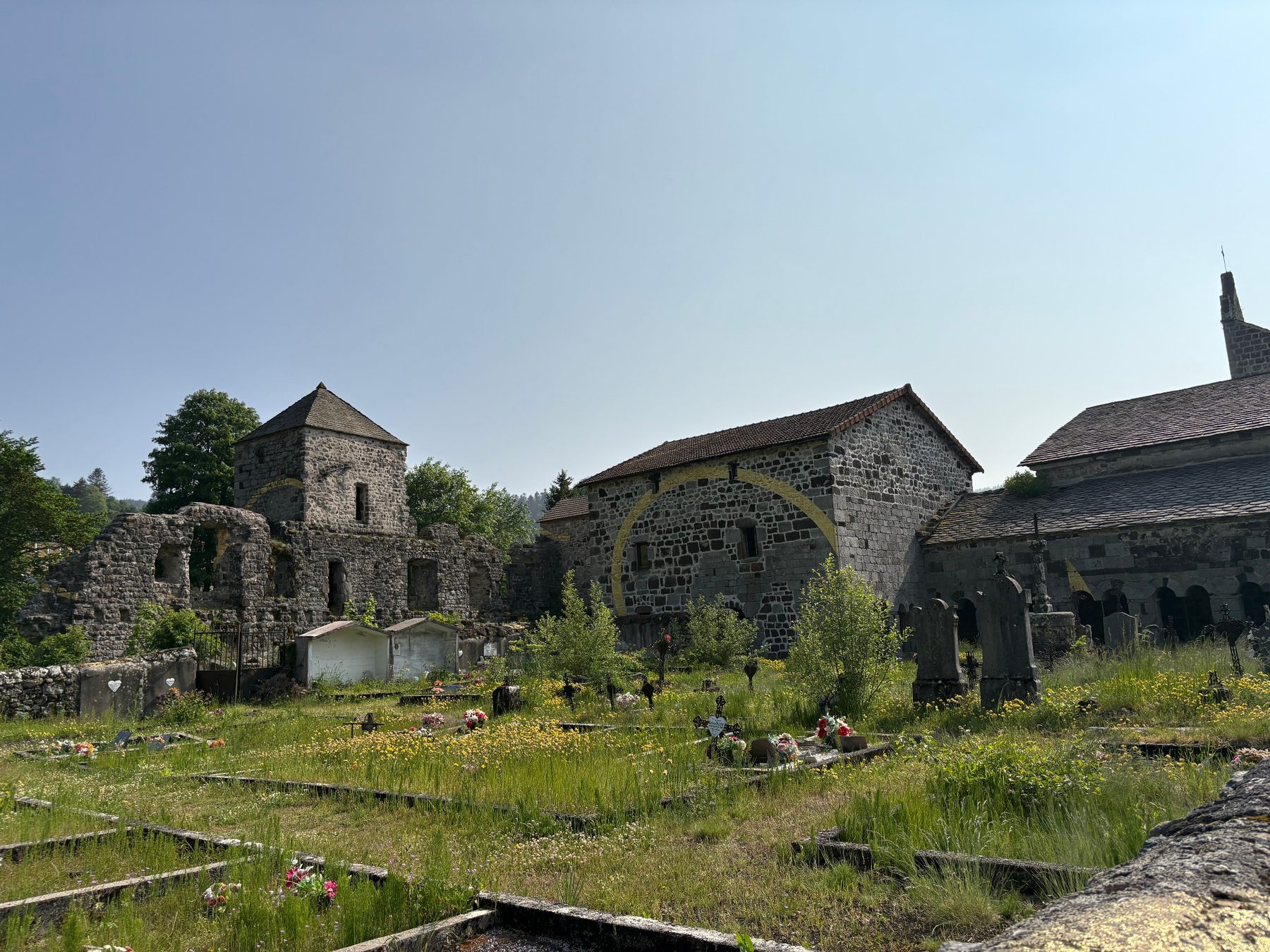 Cimetière fleuri devant une tour carrée, des vestiges en ruine et des bâtiments de pierre marqués de fragments de cercle dorés