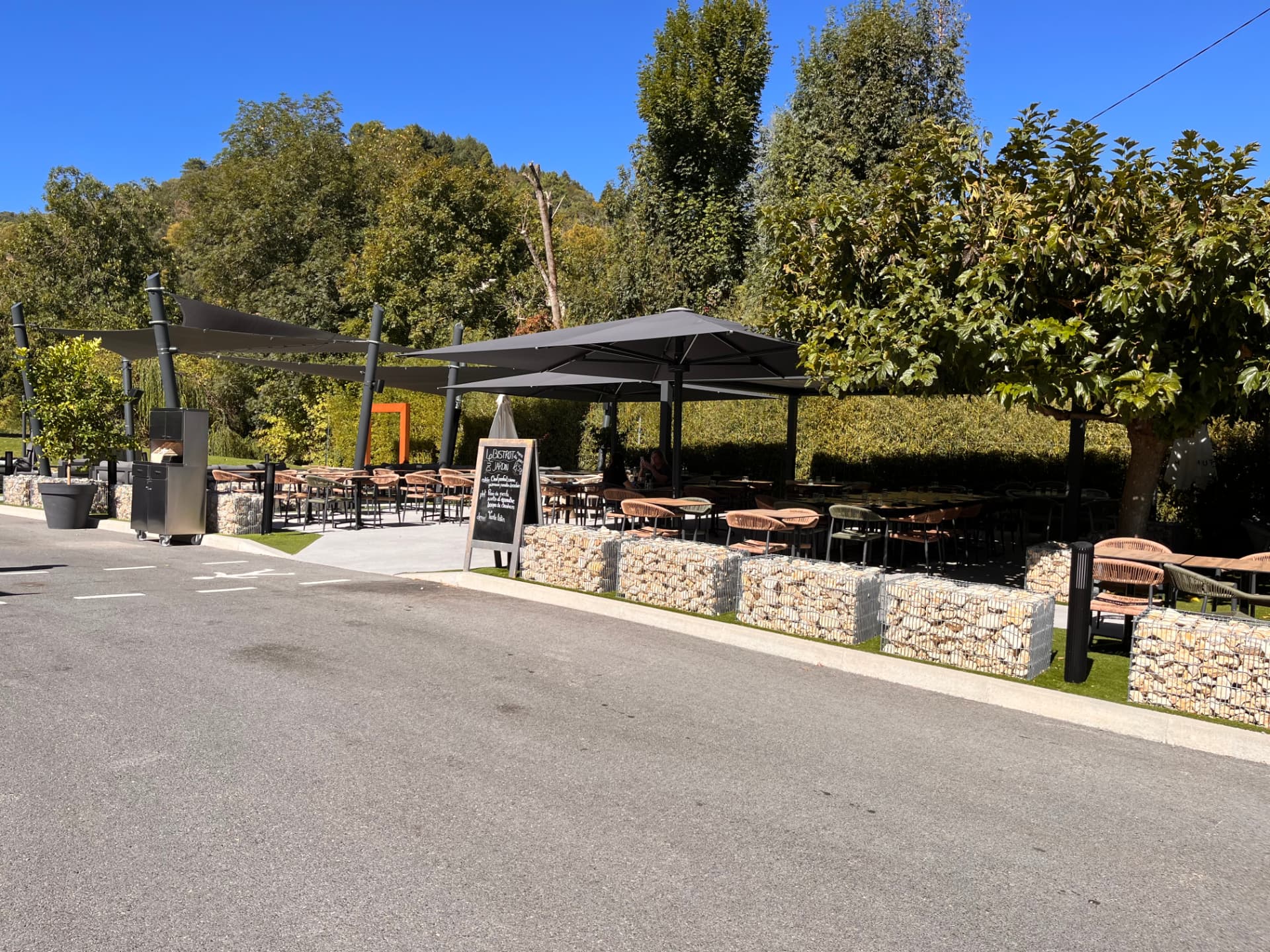 Terrasse extérieure du Bistrot du Jardin, tables et fauteuils sous de grands parasols et voiles d'ombrage