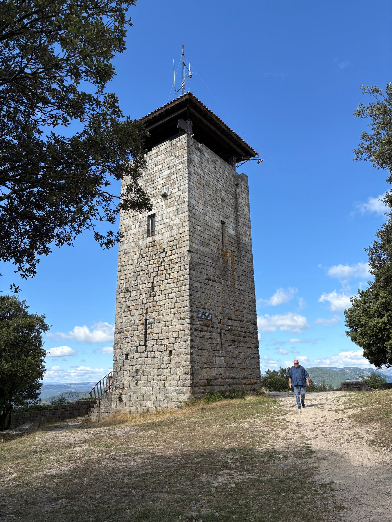 Tour de Brison, haute tour carrée en pierre coiffée d'un toit de tuiles, vue depuis le chemin d'accès sous un ciel bleu