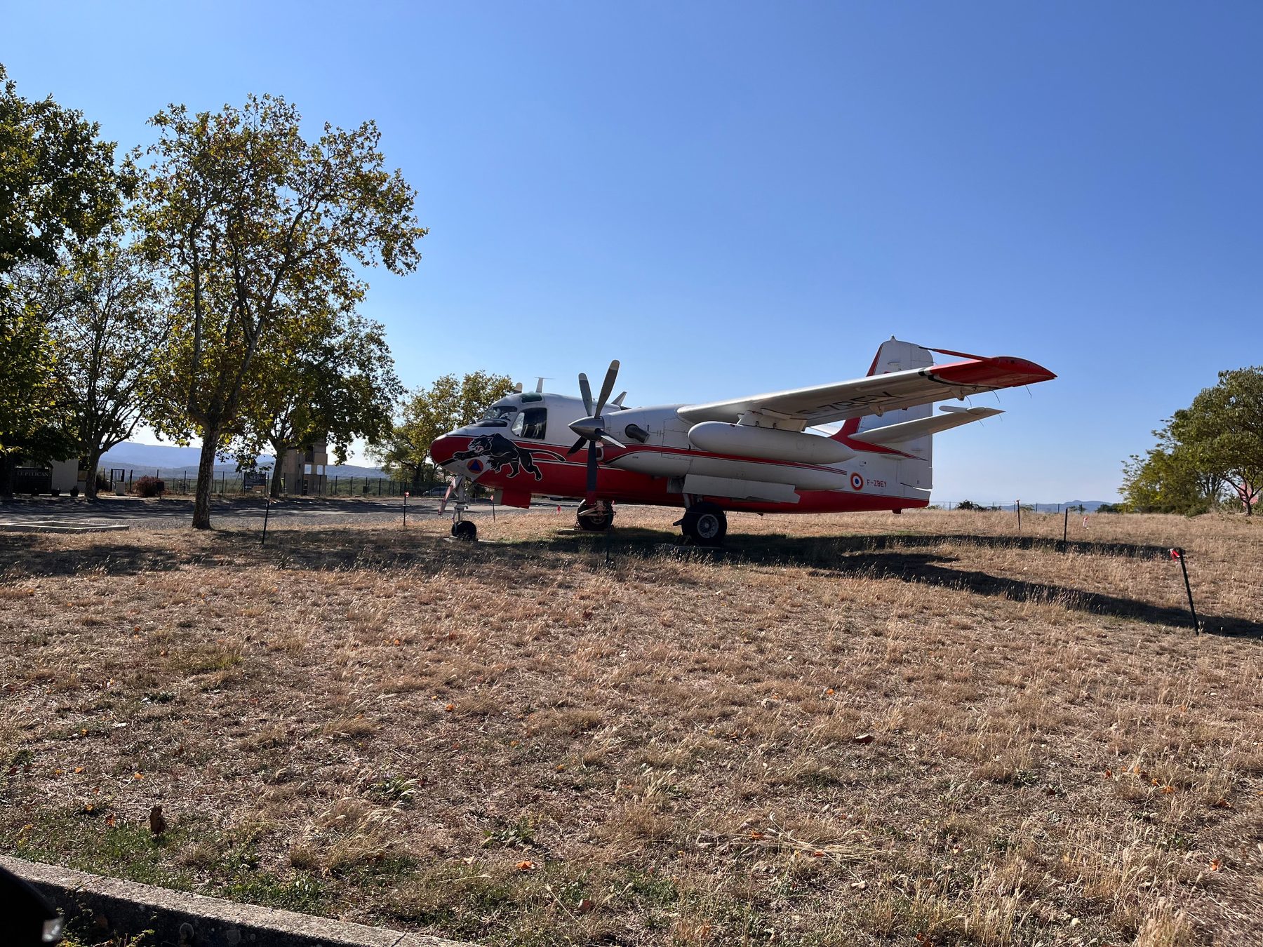 Avion bombardier d'eau rouge et blanc exposé au sol sur l'herbe sèche de l'aérodrome, sous les platanes et un ciel bleu