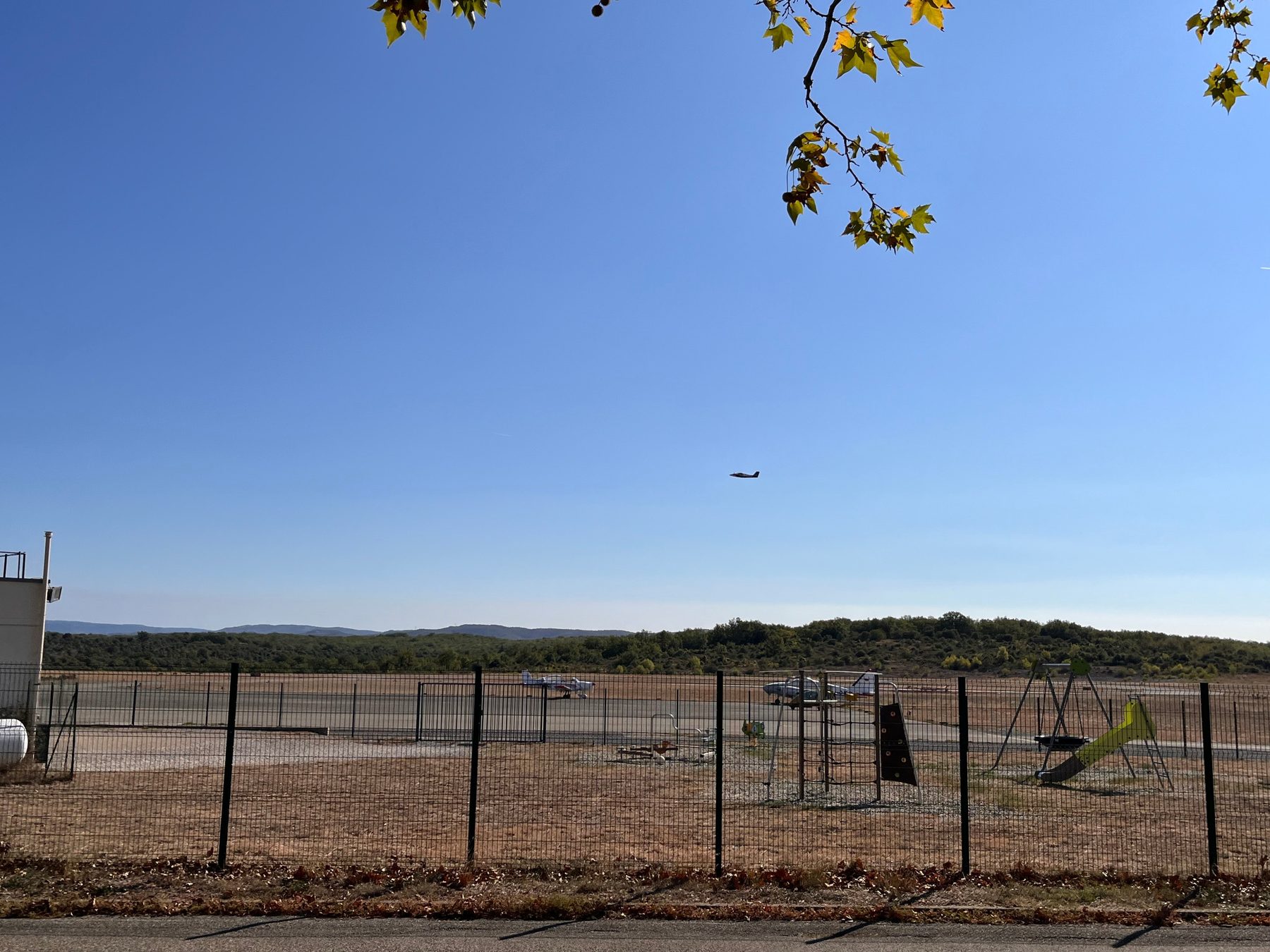 Avion en vol au-dessus de l'aérodrome, avions stationnés sur le tarmac et aire de jeux derrière la clôture