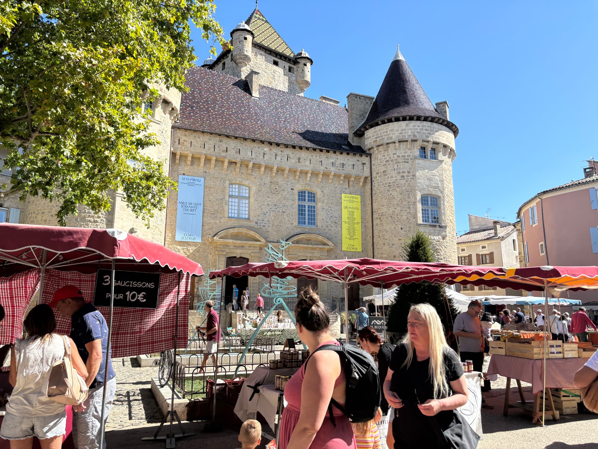 Marché d'Aubenas devant le château, étals aux auvents rouges et visiteurs sous un ciel bleu