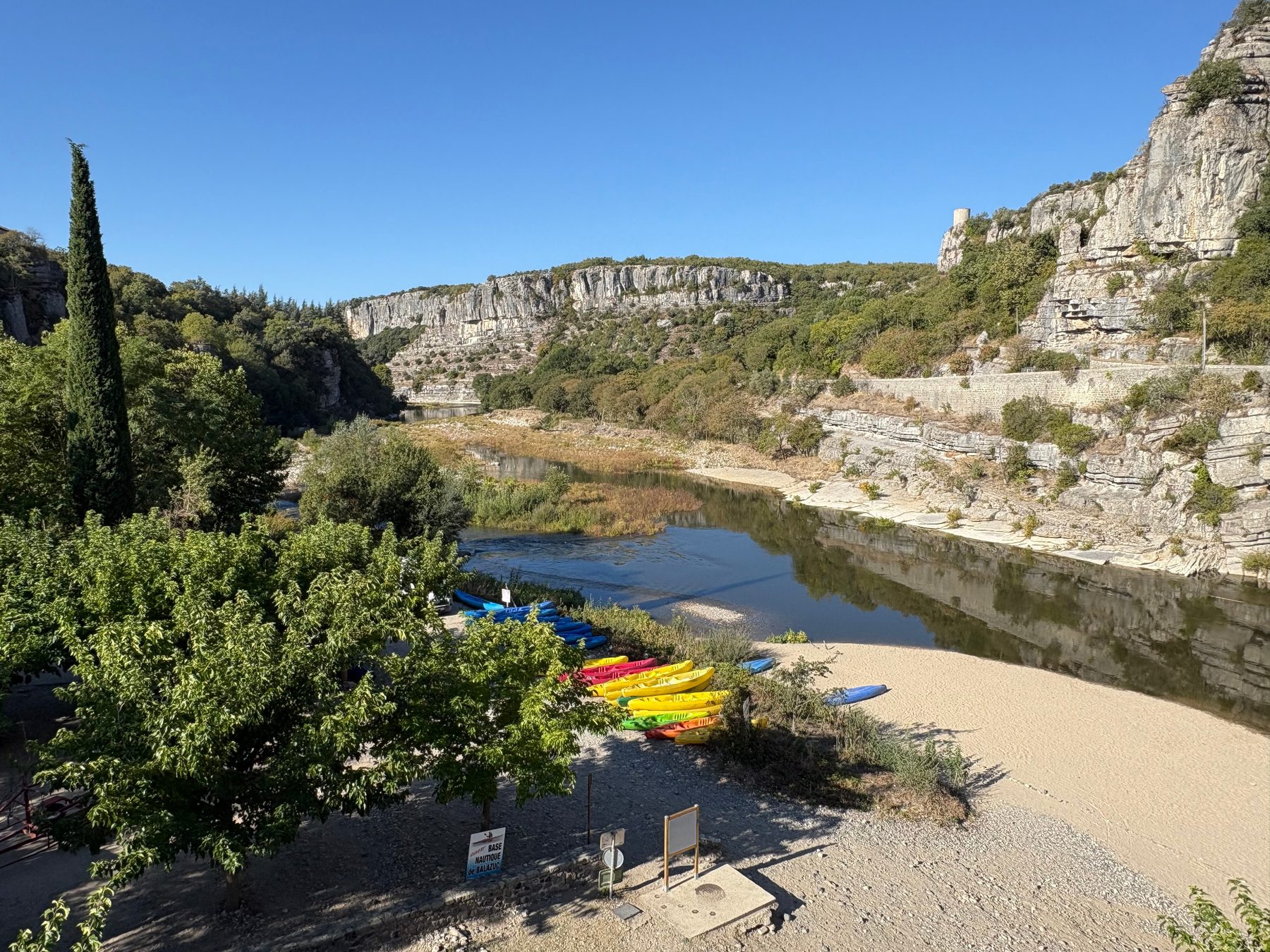 Plage de sable au bord de l'Ardèche avec des canoës colorés alignés, falaises et village en arrière-plan