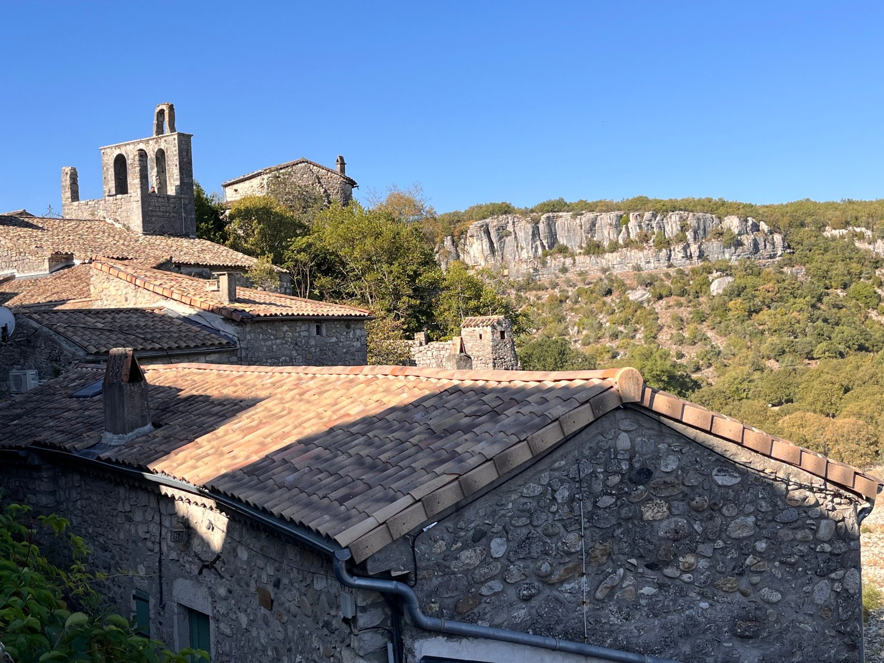 Clocher-mur ajouré de l'église dominant les toits de tuiles du village, falaises calcaires boisées en arrière-plan
