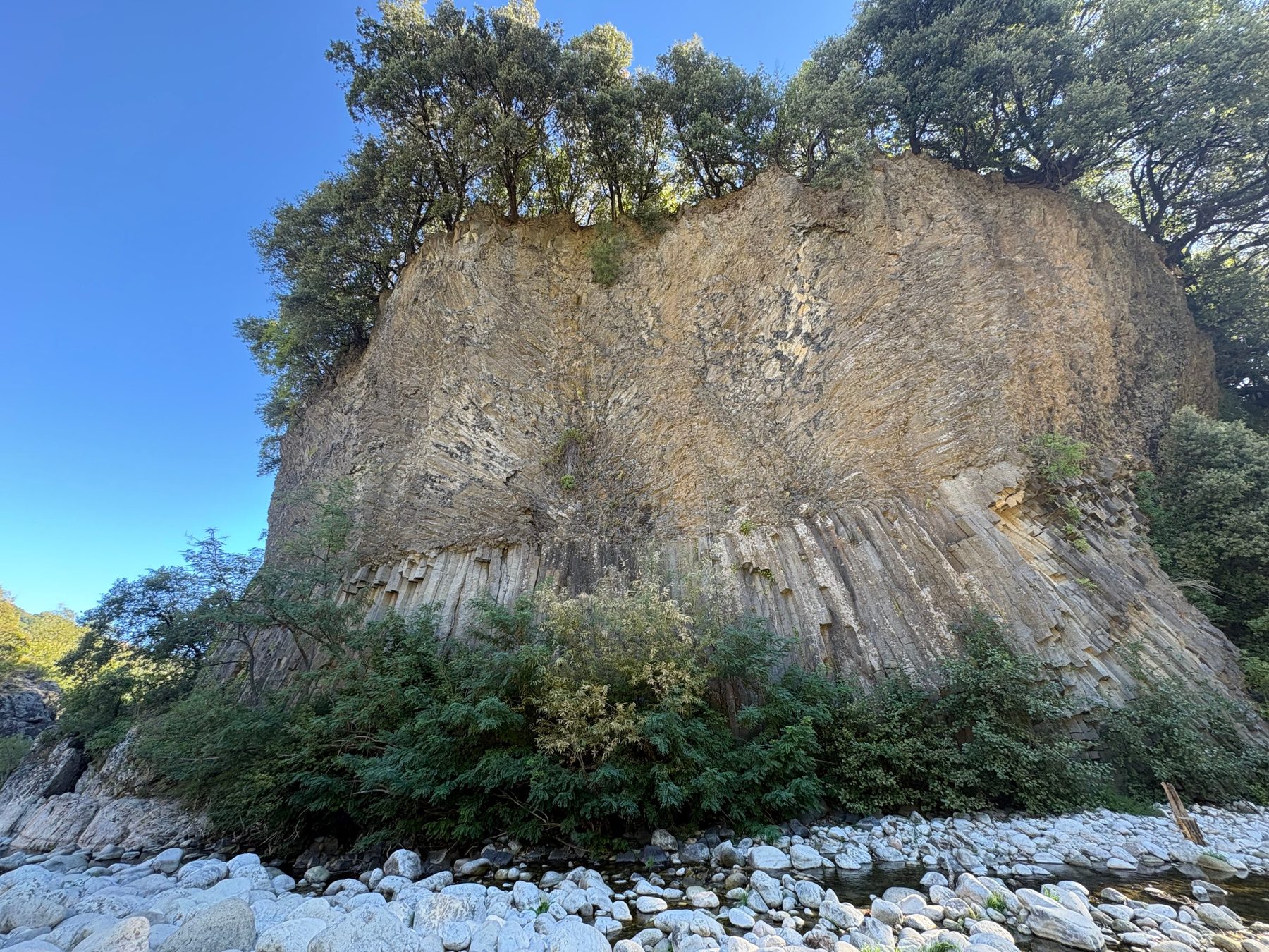 Grande paroi d’orgues basaltiques surmontée de chênes verts, au-dessus d’un lit de galets clairs sous un ciel bleu